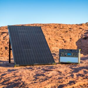פאנל סולארי קשיח BOULDER 100 בהספק 100W 12-22V ותחנת כוח ניידת ממוקמים על שטח סלעי תחת שמיים כחולים בהירים, ומציגים טכנולוגיה פוטו-וולטאית לייצור אנרגיה סולארית יעילה וממחישים תשתית אנרגיה מתחדשת ניידת בפעולה.