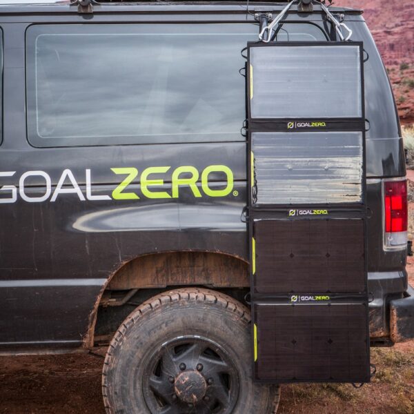 פאנלים סולאריים מתקפלים דגם 12-22V 100W NOMAD 100 של Goal Zero מוצמדים לצד רכב ואן שחור, ויוצרים תחנת ייצור חשמל סולארית ניידת בטכנולוגית פוטו-וולטאית חנייה על דרך עפר ארבעה – פתרון לאנרגיה מתחדשת מחוץ לרשת.
